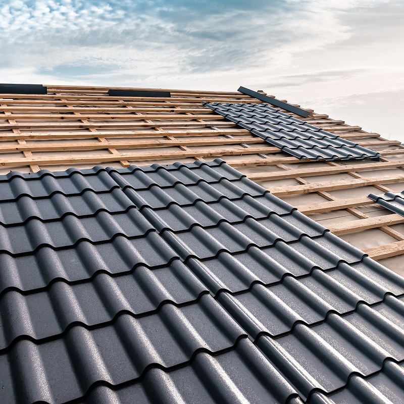 Pose de Toiture Métallique : Chantier et Installation Pose de tuiles métalliques grises sur une toiture en construction, lattes en bois apparentes sous un ciel nuageux.
