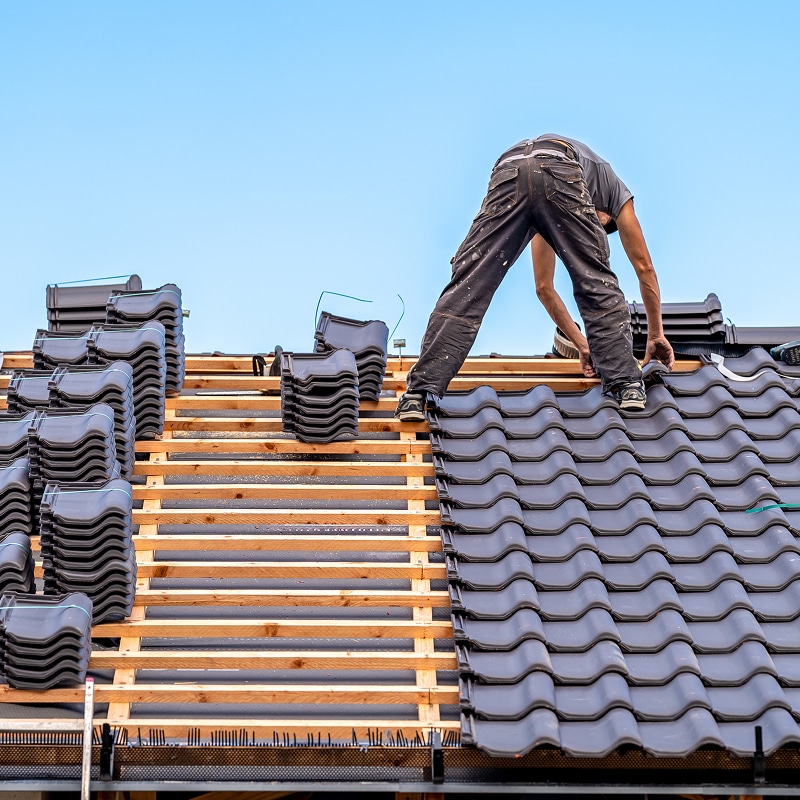 Deux couvreurs posent des tuiles sombres sur un toit en construction. Des piles de tuiles neuves attendent d'être installées.