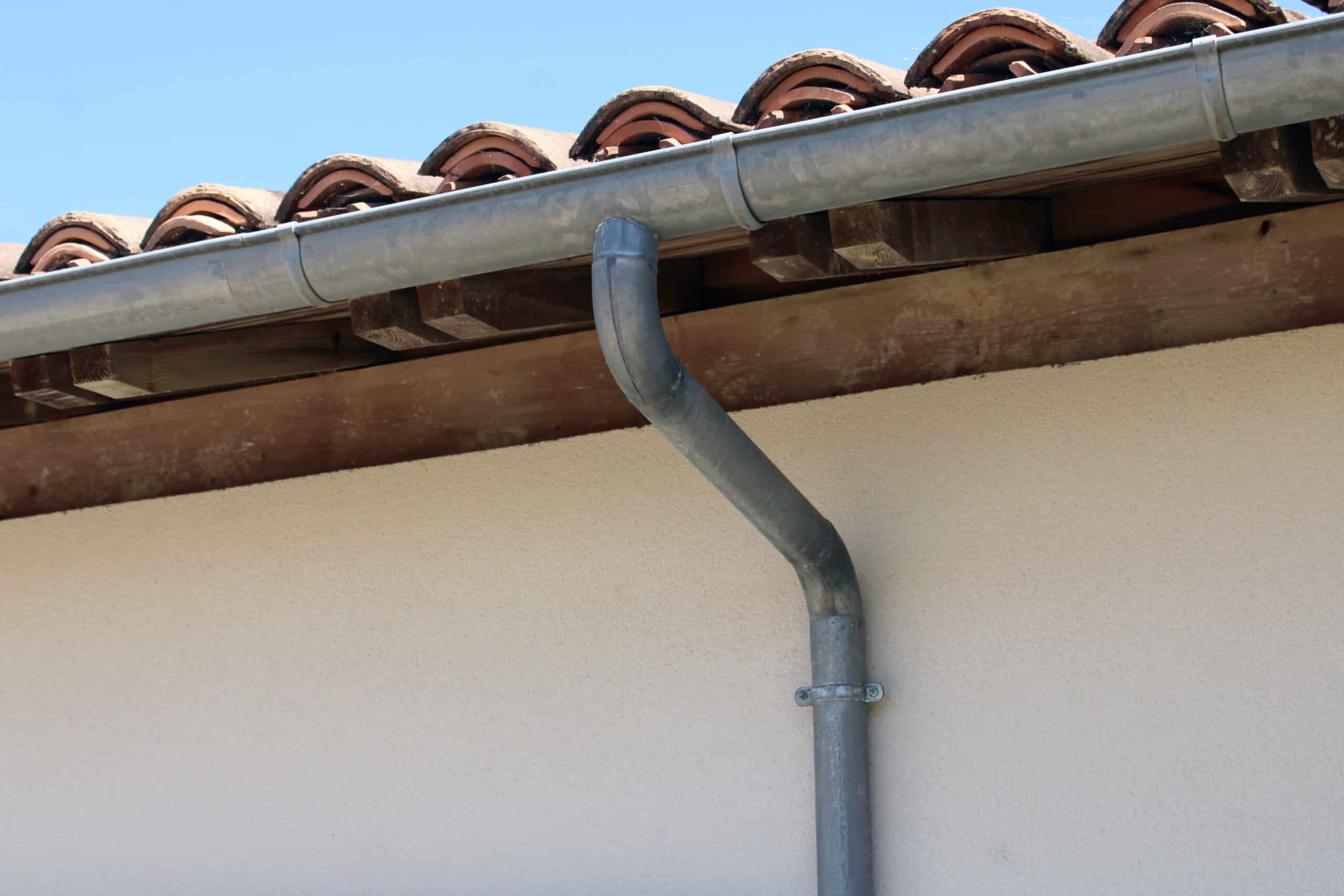 Gouttière en métal gris et tuiles de toit en terre cuite sous un ciel bleu clair, fixées à la façade beige d'une maison.
