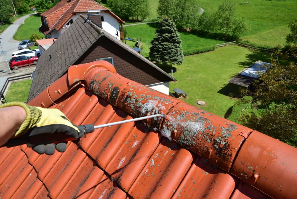 Main gantée nettoyant un toit de tuiles rouges couvert de mousse et lichen avec un pulvérisateur, fond de maisons et verdure.