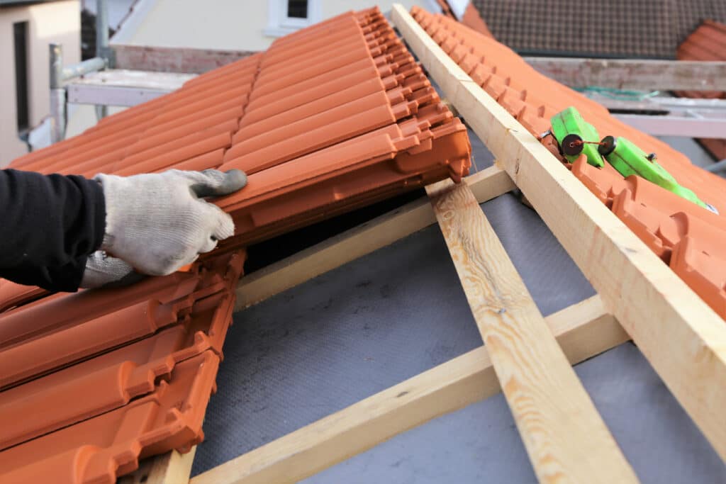 Main gantée posant des tuiles en terre cuite sur un toit en construction, avec chevrons en bois et membrane d'étanchéité visibles.