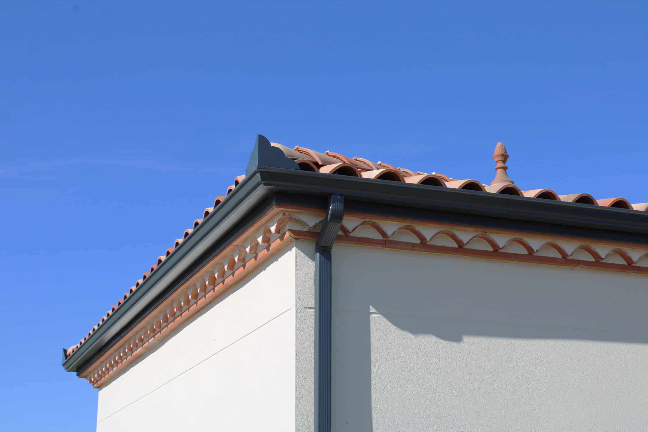 Coin de maison méditerranéenne avec toit de tuiles ocres, gouttière anthracite, frise décorative et mur blanc sous ciel bleu.