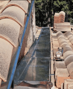Installation de gouttière en zinc entre un mur ancien et des tuiles de toit en terre cuite. Une meuleuse et des tuiles neuves sont sur la toiture en rénovation.