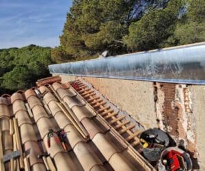 Rénovation de toiture avec tuiles en terre cuite et gouttière métallique neuve. Outils de chantier sur les lattes de bois, mur de briques exposé. Forêt en arrière-plan.